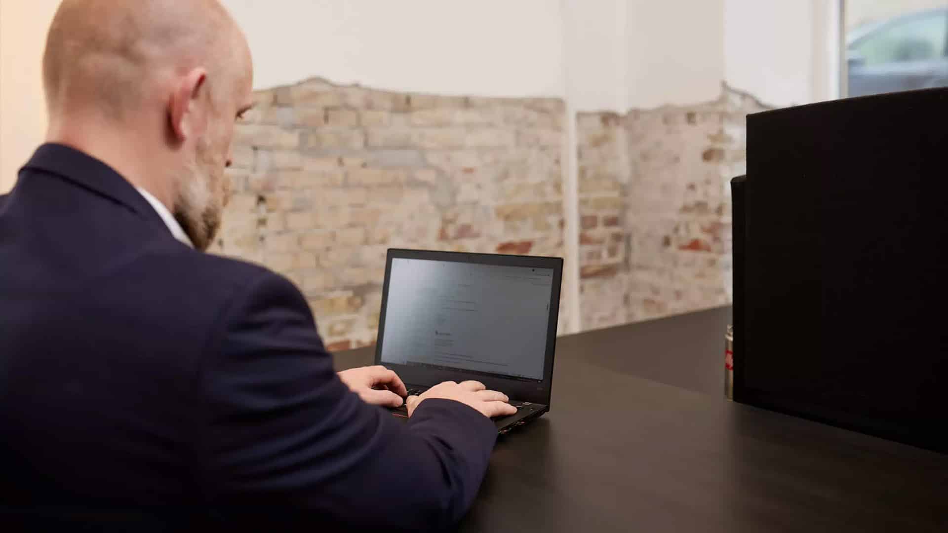 Mand i jakkesæt arbejder på en bærbar computer ved et skrivebord i et kontormiljø.