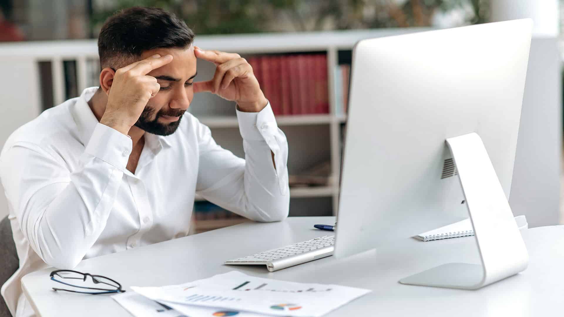 Mand med kort hår og skæg sidder ved skrivebord og holder hovedet med hænderne foran en computer.